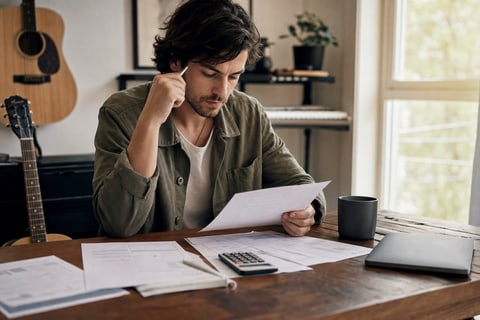 Editorial photograph of a musician at a home desk reviewing a document and financial figures with a calculator and laptop nearby, natural window light, acoustic guitar visible in the background, no readable text on documents or screen.