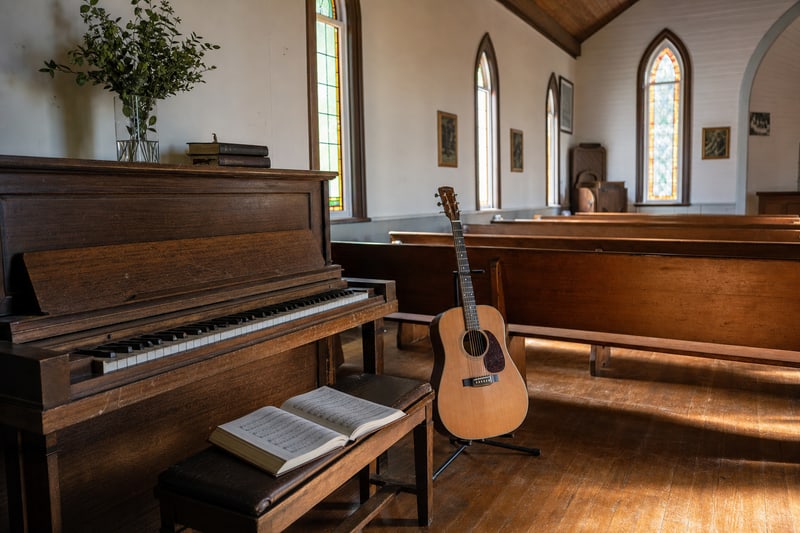 Photograph of a small country church interior with an upright piano, an open hymnal on the bench, an acoustic guitar leaning against the end of a pew, tall arched stained-glass windows, and warm wood floors.