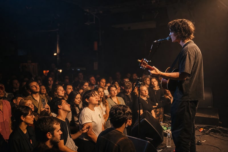 Editorial photograph of a developing artist performing on stage at a small intimate venue with an engaged crowd visible in the foreground, warm stage lighting, artist-audience connection clear, no arena scale, no readable logos.