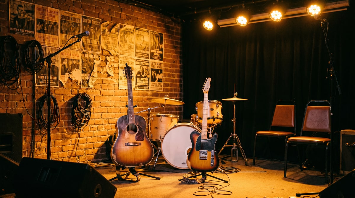 Editorial photograph of an independent country band performing on a small stage in a dimly lit venue with attentive seated audience and a guitar case open in the foreground.