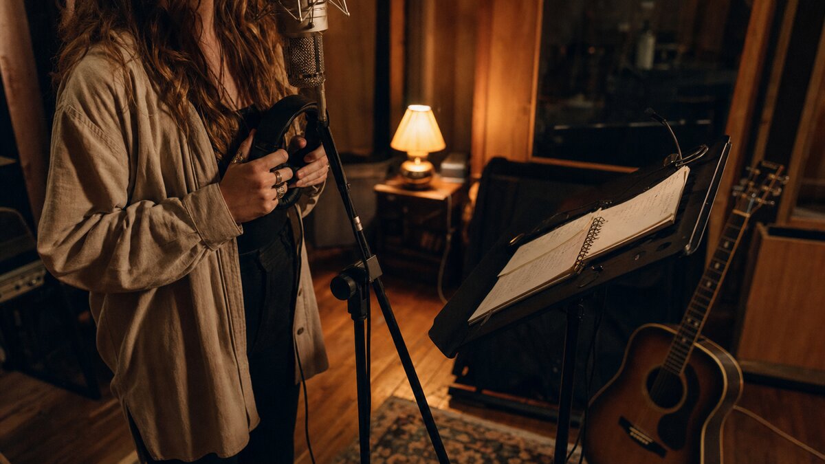 A vocalist in a small warm-lit studio at a vintage-style microphone with headphones on, an acoustic guitar resting nearby, and handwritten lyric drafts on a music stand.