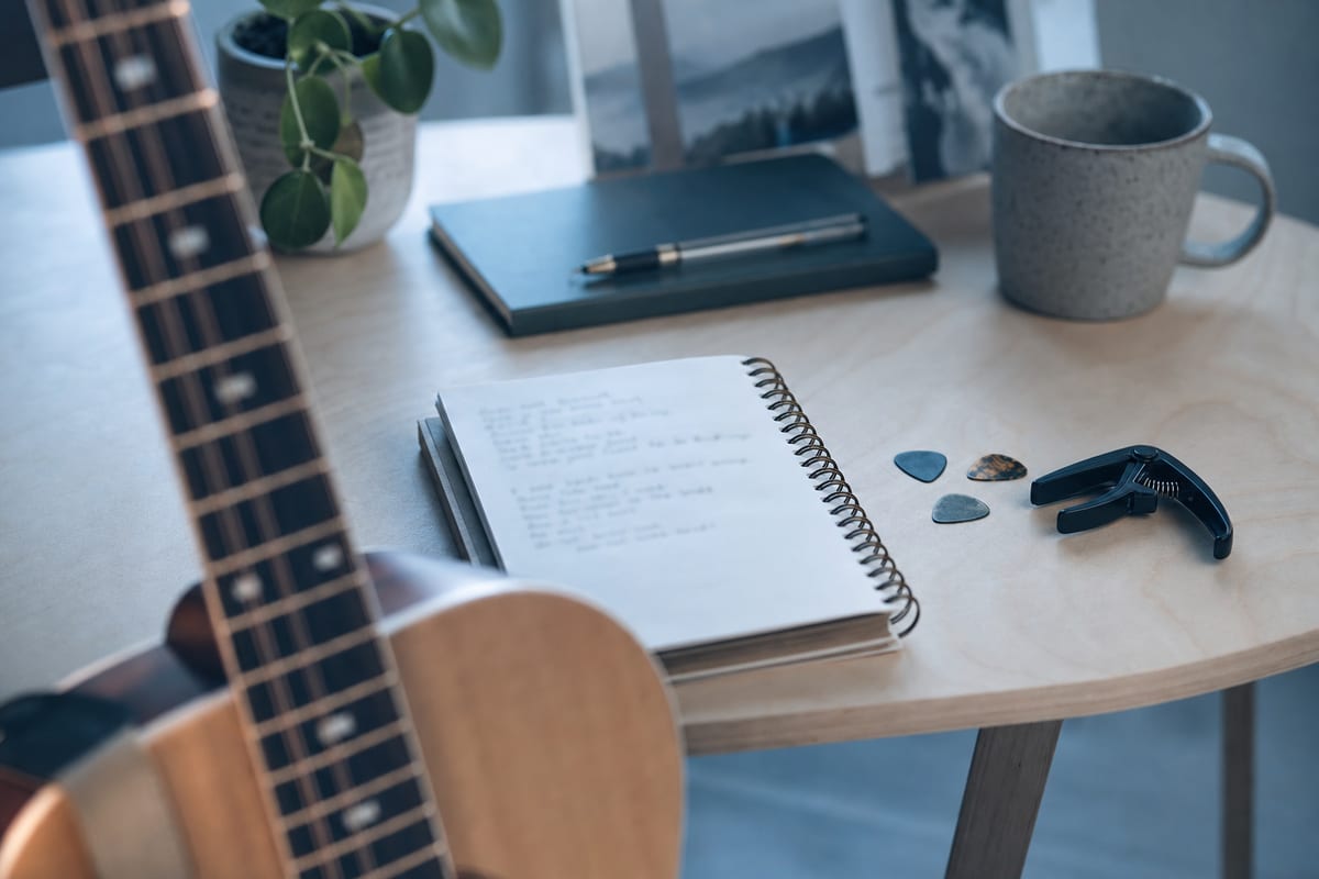 Editorial photo: a songwriter's notebook with handwritten lyric sections next to an acoustic guitar on a wooden floor, soft natural light, no readable text